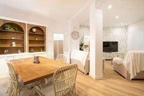 A modern interior with a wooden dining table, white wicker chairs, a sofa, a TV, and a clock on the wall.