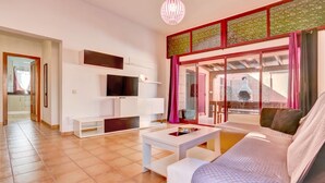 A modern living room with a sofa, a coffee table, a flat-screen TV, and a large window with curtains. - Villa Malva: Private Jacuzzi, Ping Pong & Relaxation in Corralejo (Corralejo)