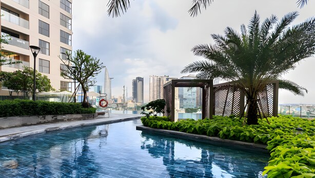 A swimming pool with tiled flooring, surrounded by greenery and palm trees, with high-rise buildings in the background.