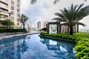 A swimming pool with tiled flooring, surrounded by greenery and palm trees, with high-rise buildings in the background. - Luxury Millenium Apartment (Ho Chi Minh City)