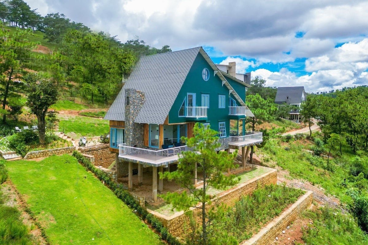A large, two-story house with a balcony, surrounded by a lush green landscape and hills.
