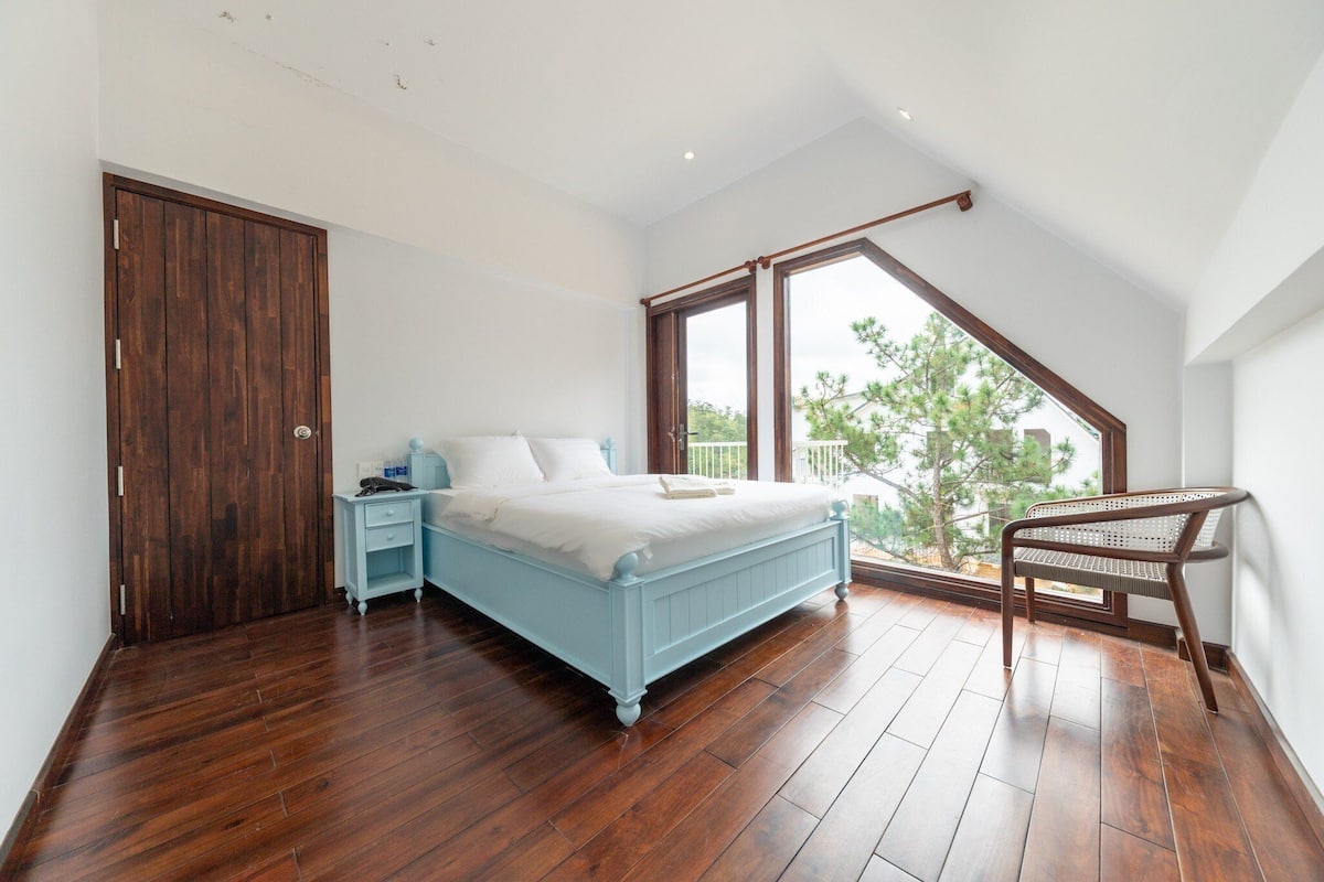 A bedroom with a wooden floor, a bed with white bedding, a nightstand, a wooden wardrobe, and a window with a view of trees.