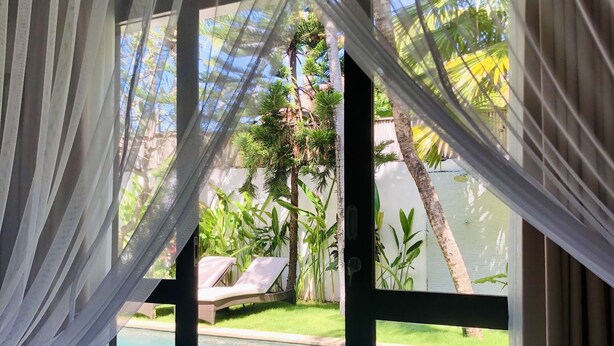 Ein Zimmer mit Bett, Blick auf einen Swimmingpool und einem Balkon mit Pflanzen und einem Stuhl.