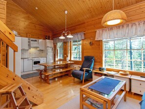 Ein gemütliches Blockhauss-Interieur mit Holzvertäfelung, einem Essbereich, einer Küche mit weißen Geräten und einem großen Fenster mit Blick auf den Wald.