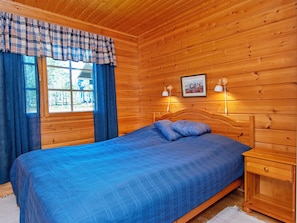 A wooden cabin bedroom with a bed, curtains, a window, and a wall-mounted lamp.
