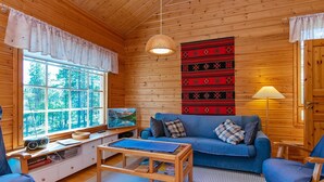 Un acogedor interior de cabaña con paredes de madera, un sofá azul, una mesa de centro de madera, un televisor y un ventanal con vistas a árboles.