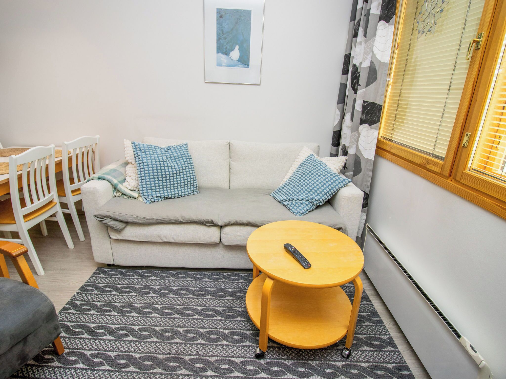 Un salon avec un canapé, deux coussins bleus, une table basse ronde en bois, une télécommande, un tapis à motifs et une fenêtre avec des stores.