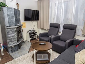 Un salon doté d’une cheminée en pierre, d’un téléviseur à écran plat, de deux fauteuils gris, d’une table basse ronde et d’un canapé agrémenté d’un coussin rouge.