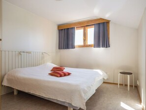 Une chambre à coucher avec un lit, une fenêtre avec des rideaux, un radiateur et un petit tabouret.