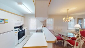 A modern kitchen with white cabinets, a microwave, and a coffee maker. There is a dining area with a glass table and wicker chairs.