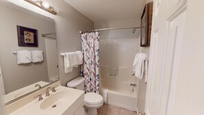 Badeværelse med håndvask, toilet og badekar. Der er håndklæder hængende på væggen og en bruseforhæng.