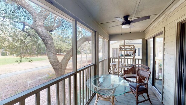 A covered porch with a glass table and wicker chairs, a ceiling fan, and a view of a tree and a building in the background. - Pawleys Plantation Greens 1 Bedroom Retreat (Pawleys Island)