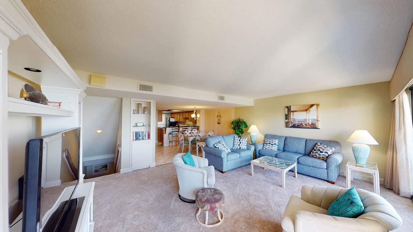 A living room with a sofa, armchairs, a coffee table, and a TV. There is a kitchen in the background.