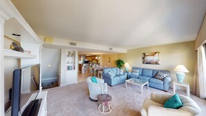 A living room with a sofa, armchairs, a coffee table, and a TV. There is a kitchen in the background. - Coastal Hideaway 2 Bedroom Condo with Ocean Views (Pawleys Island)