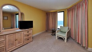Habitació d'hotel amb un mirall gran, una televisió sobre un moble de fusta, una cadira de vimet i vistes a l'oceà a través d'una finestra amb cortines rayades.