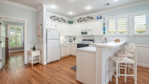 A kitchen with white cabinets, a central island, and a dining area with white chairs. - Beautiful 2BR 2BA Lakeside Villa with Lake Views (Pawleys Island)