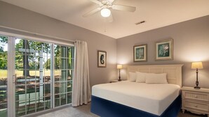 Une chambre dotée d’un grand lit, d’un ventilateur de plafond, d’une table de chevet avec une lampe, et offrant une vue sur un espace extérieur grâce à une porte-fenêtre coulissante.
