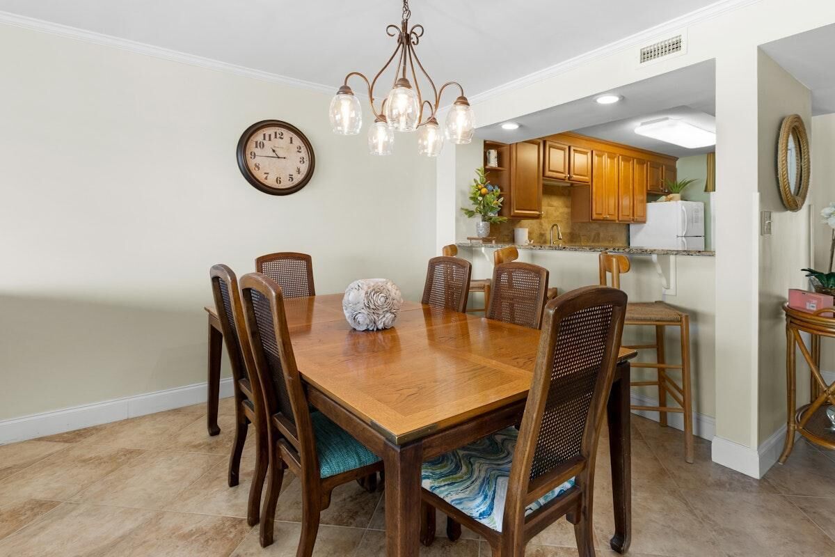 A dining room with a wooden table and chairs, a chandelier, a clock, and a kitchen in the background.