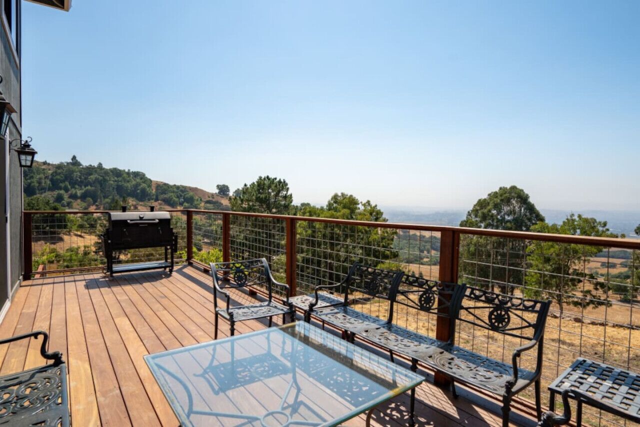 A wooden deck with metal chairs and a glass table, overlooking a hilly landscape.