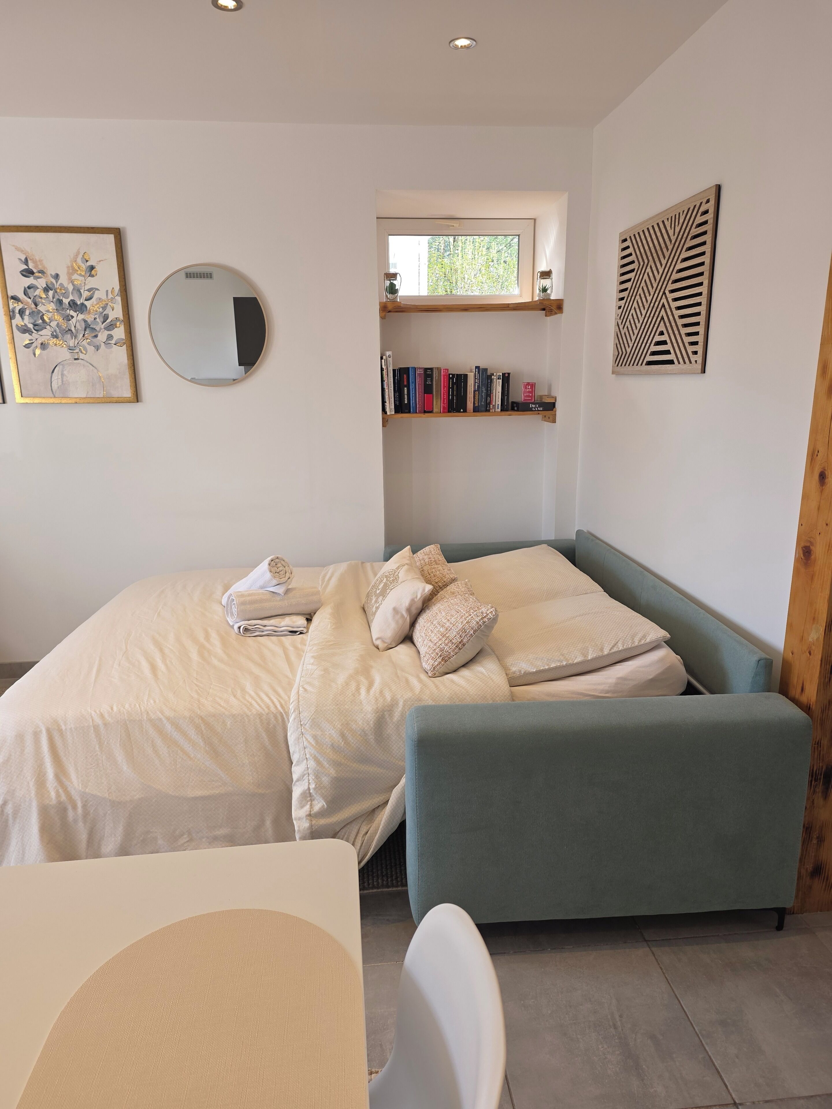 Une chambre à coucher comprenant un lit, une table de chevet, une chaise et une fenêtre surmontée d’étagères.