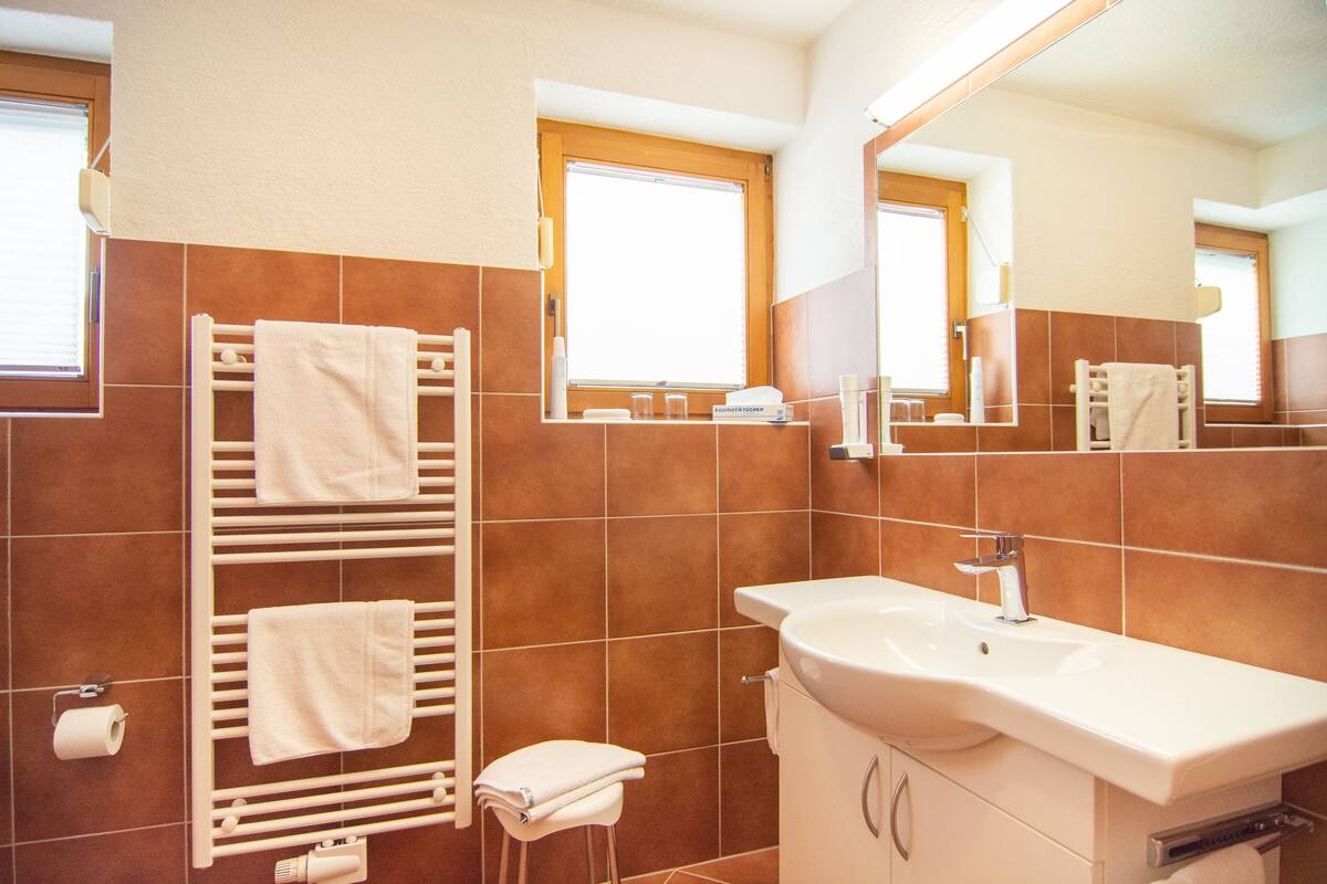 A bathroom with a white sink, a towel rack with multiple towels, a toilet, and a window with blinds.