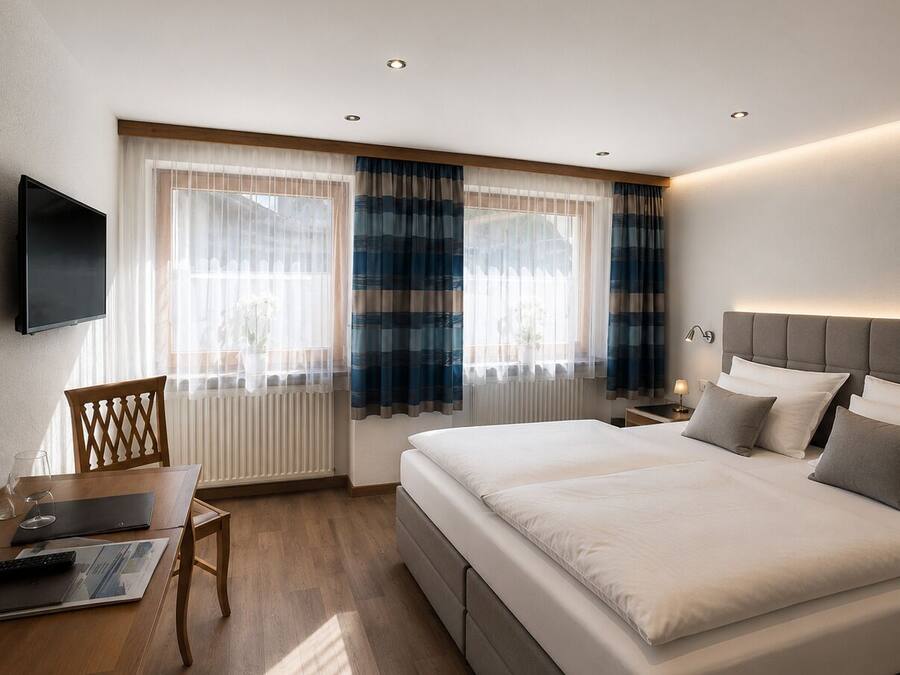 A modern hotel room with a large bed, a desk with a chair, a flat-screen TV, and a window with curtains.