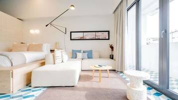 A modern hotel room with a bed, sofa, and a small table. There is a large window providing a view of the outside.