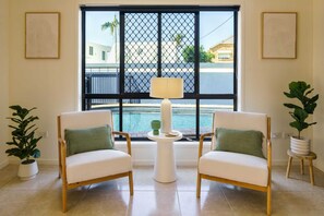 A room with two white chairs, a small white table with a lamp, and a window with a view of a pool and buildings.
