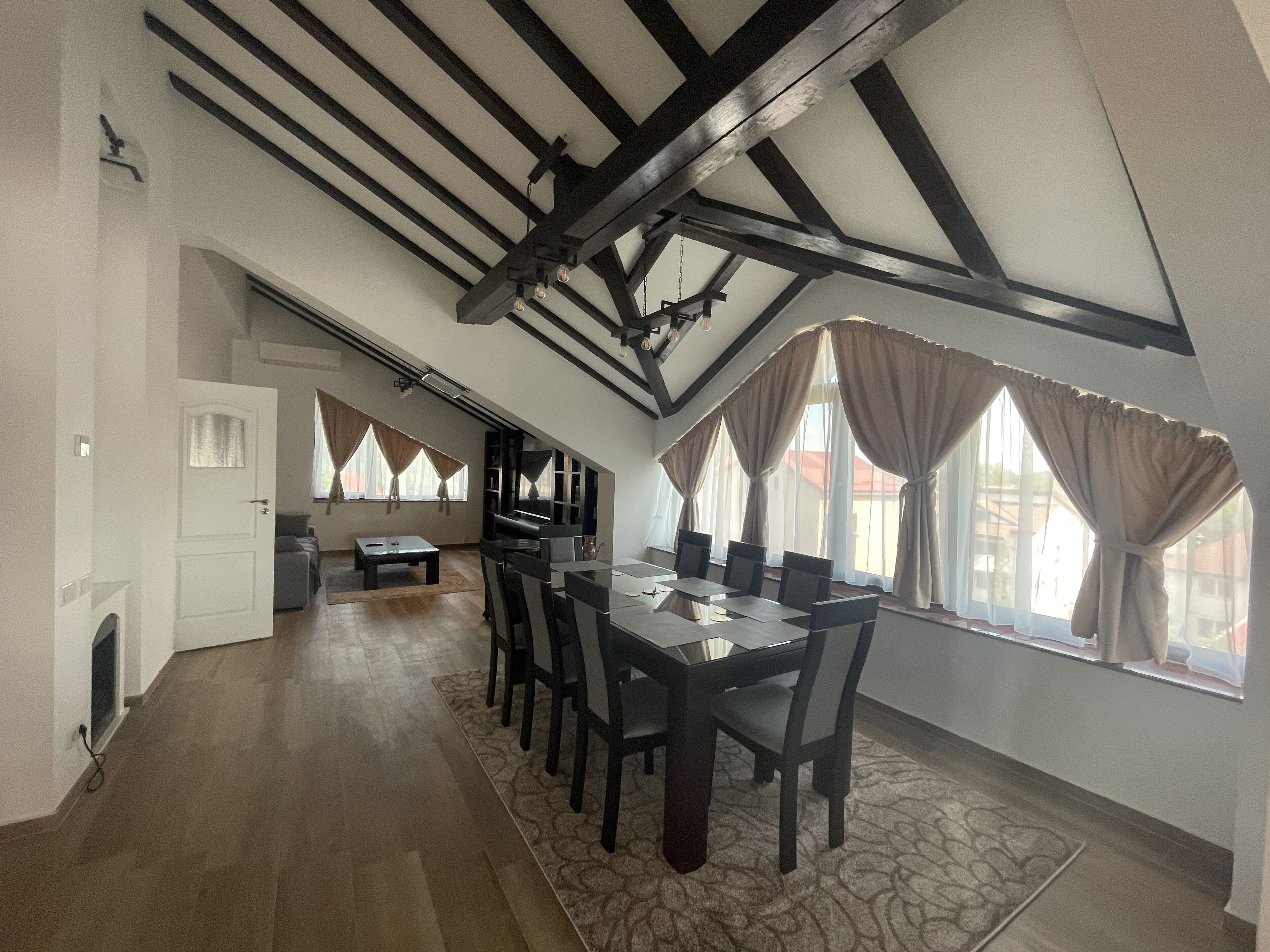 A spacious dining area with a high ceiling and exposed beams, a large table with chairs, and a view of the outside through large windows with curtains.