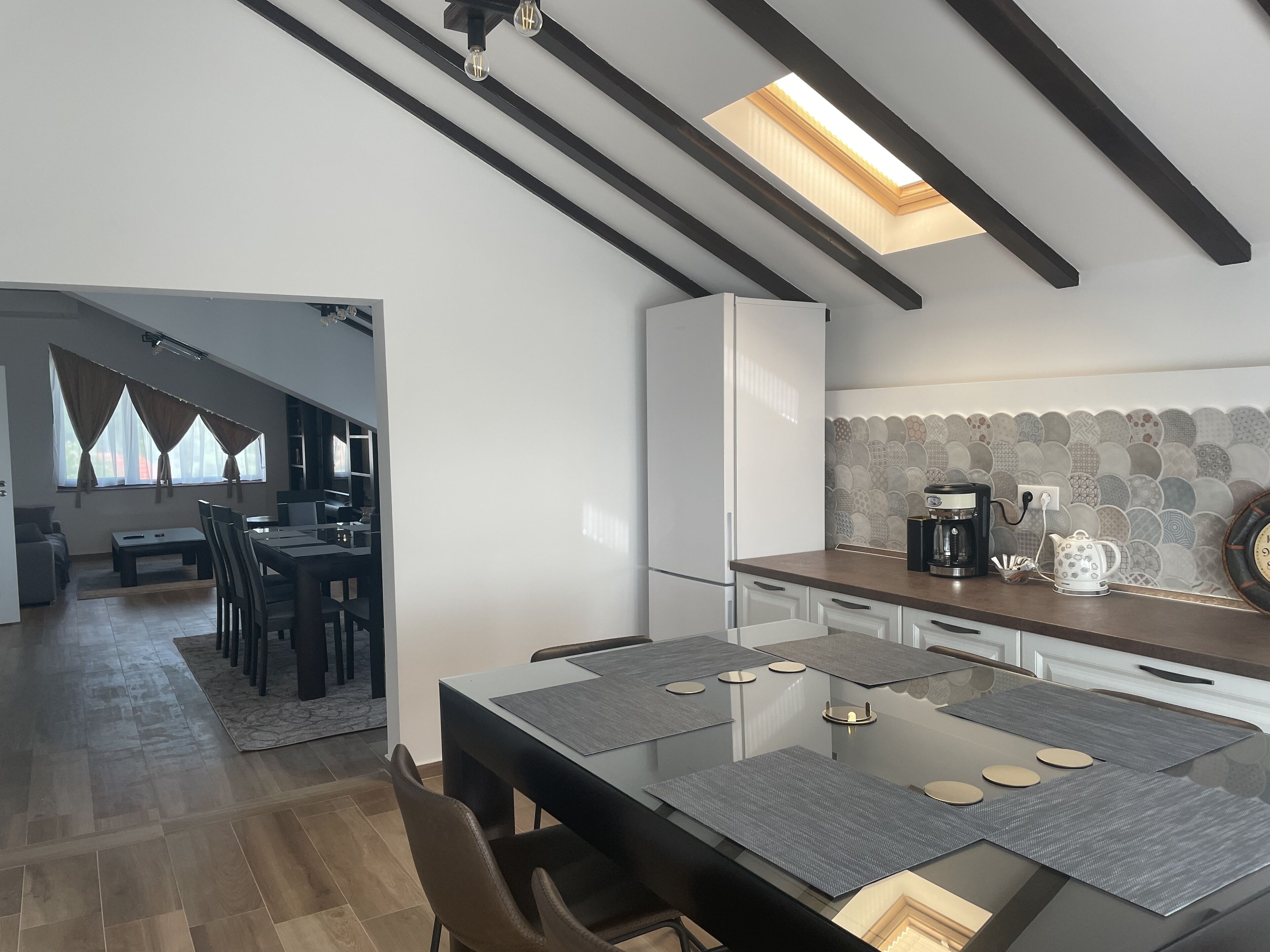 A modern kitchen with a dining area, featuring a skylight, a coffee maker, and a patterned backsplash.