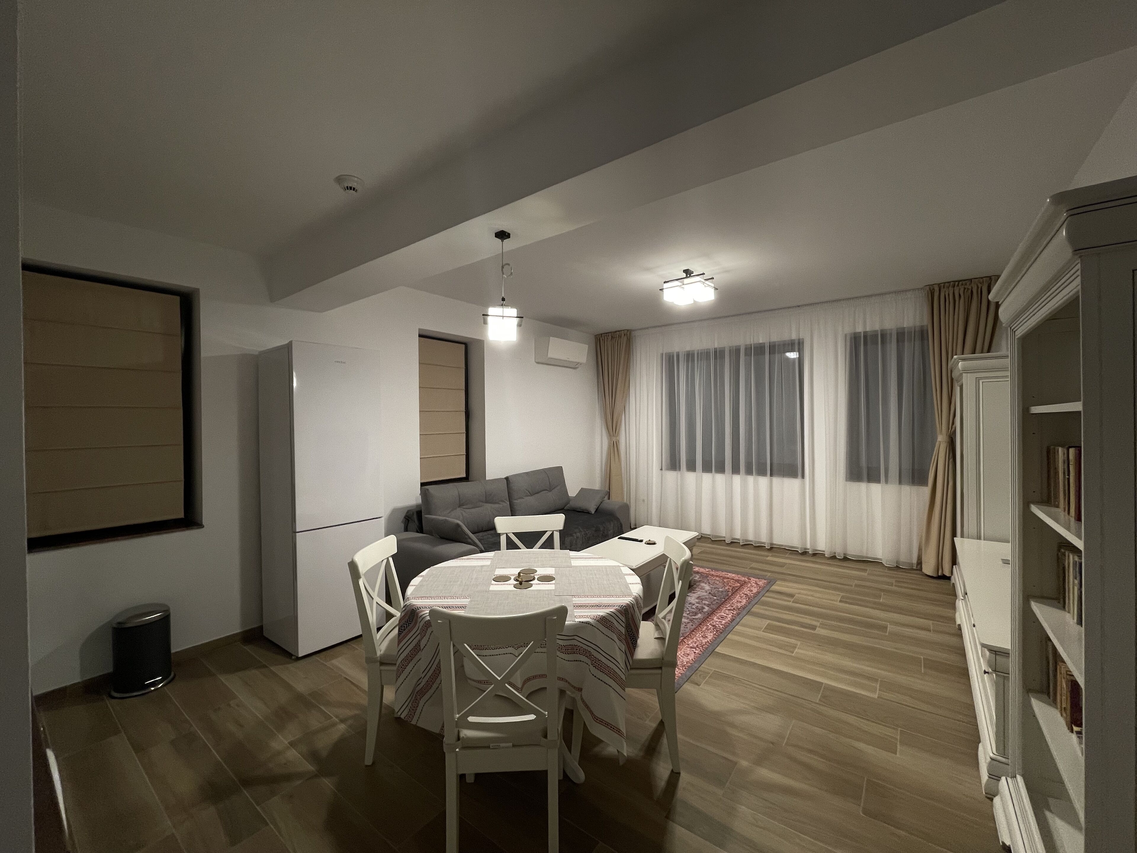 A modern living room with a sofa, dining table, and chairs. There is a bookshelf and a window with curtains.