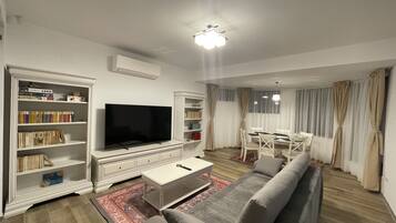 A living room with a flat-screen TV, a bookshelf, a sofa, and a dining area with a table and chairs.