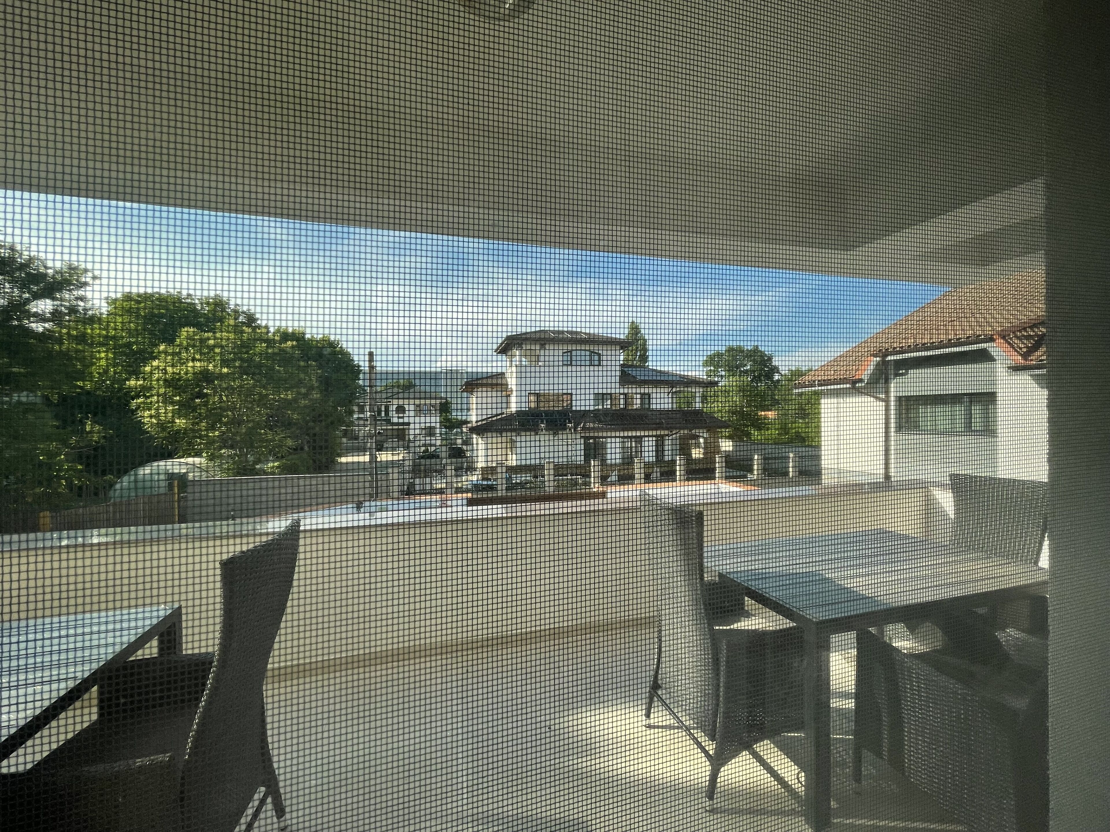 Une vue à travers une fenêtre munie d’un grillage métallique, qui révèle un bâtiment à la façade blanche et au toit recouvert de tuiles rouges, une zone pavée avec des chaises et des tables, et des arbres en arrière-plan.