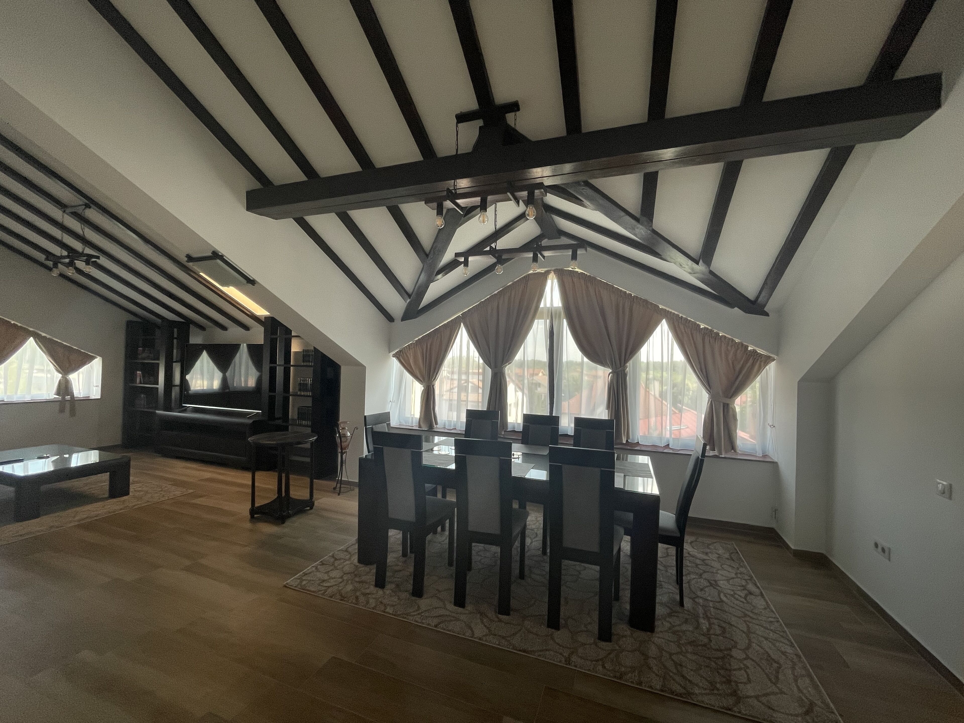 A spacious loft with exposed wooden beams, a dining area with a large table and chairs, and a cozy seating area with a sofa and a TV.