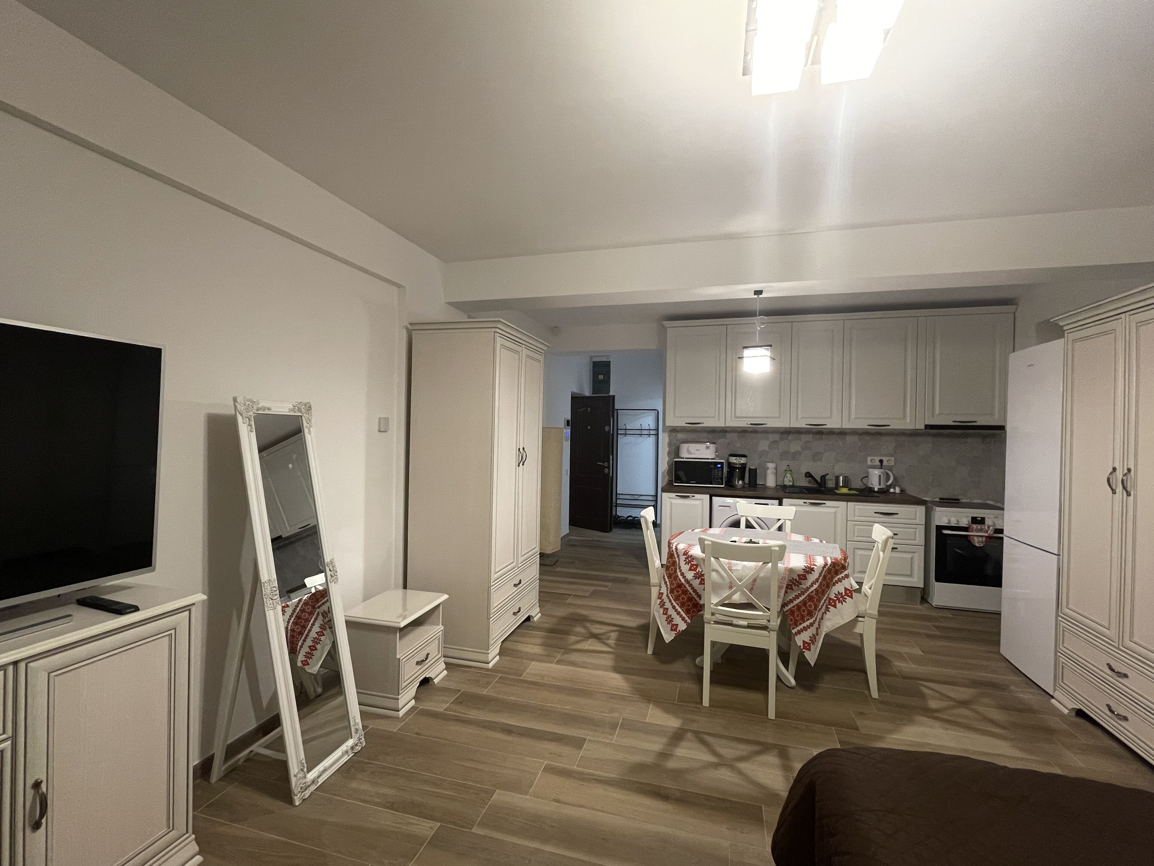 A compact kitchen with white cabinets, a dining table with chairs, a TV, and a large mirror.