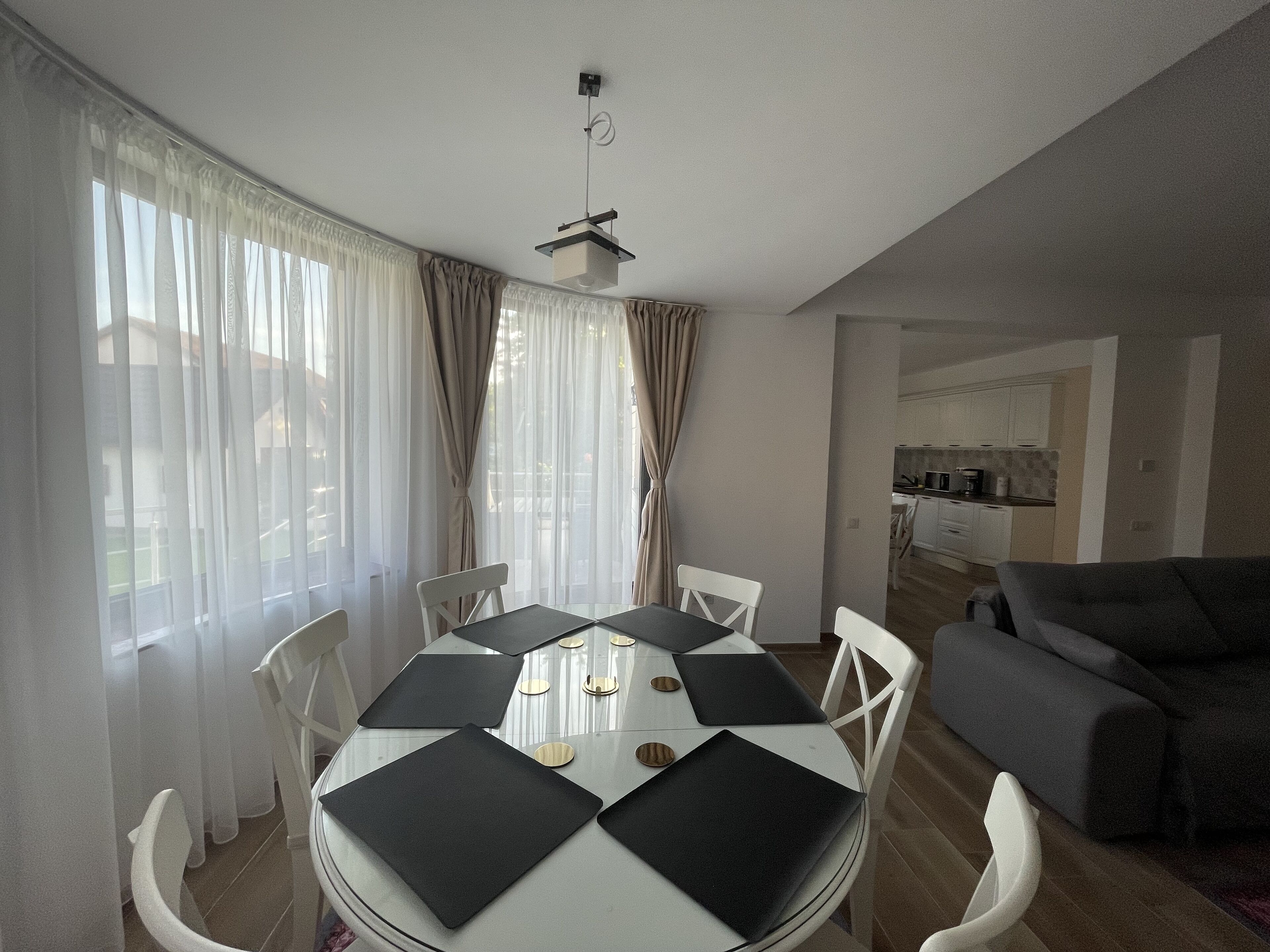 Comedor con una mesa redonda, sillas blancas y manteles individuales negros. Hay un sofá gris y una cocina al fondo.