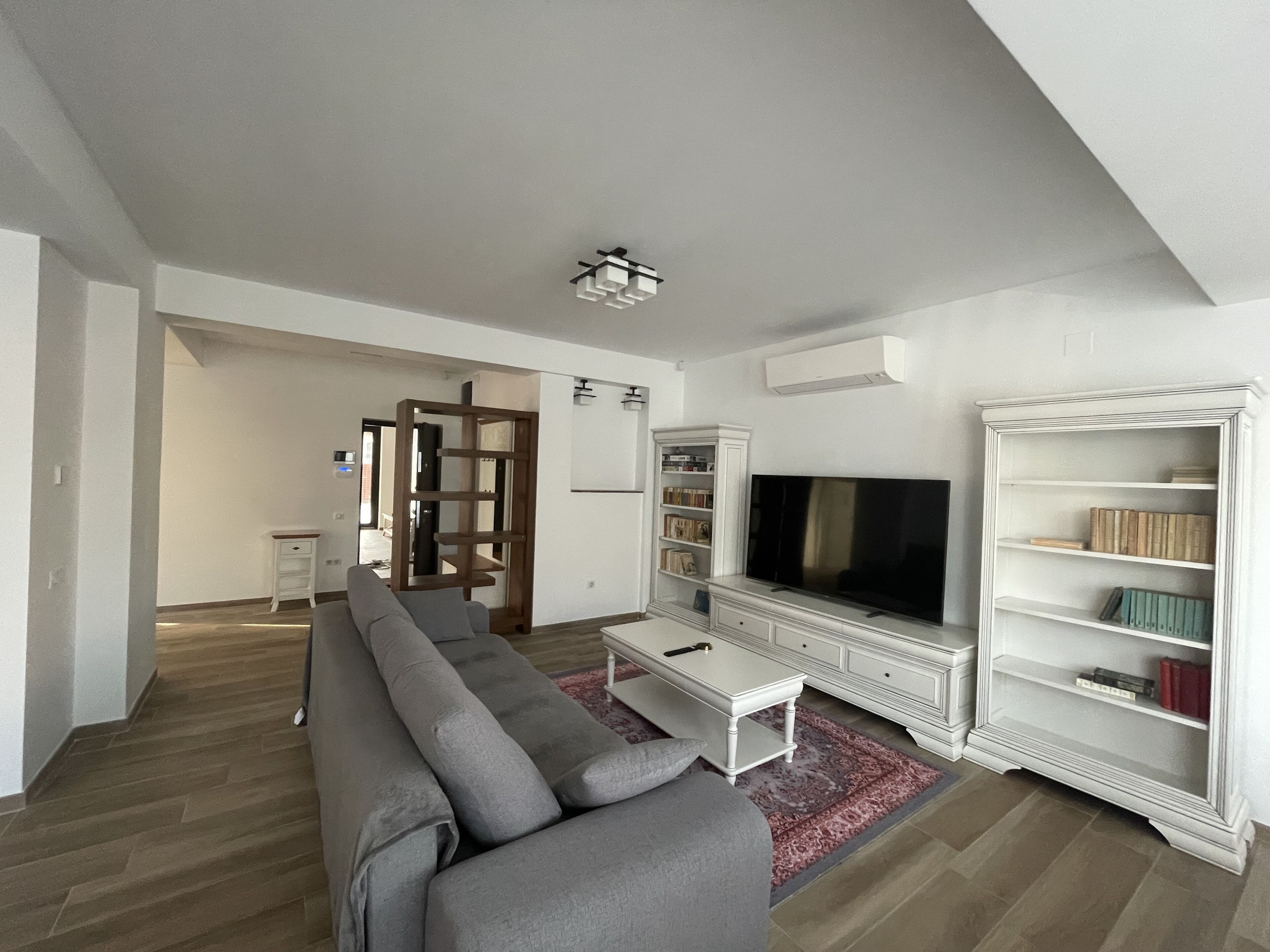 A modern living room with a grey sofa, a flat-screen TV, a white entertainment unit, and a bookshelf.