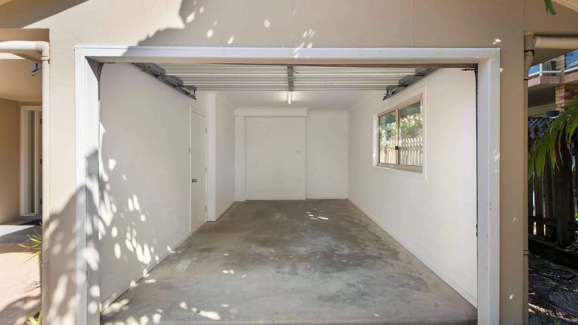 Spazio ristretto e chiuso, con pavimento in cemento e pareti bianche, dotato di una porta e una finestra che si affaccia su una recinzione e degli alberi.