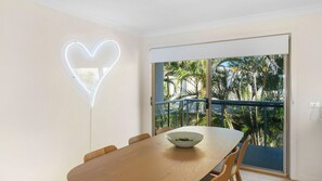 Un coin repas avec une table en bois, des chaises, un luminaire en forme de cœur et une vue sur les palmiers, visible à travers une porte-fenêtre coulissante.