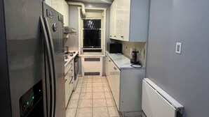 A narrow kitchen with a refrigerator, stove, and microwave. There is a window with a curtain, and a radiator on the wall. - Two Large Cozy Bedroom Condominium that is 20 mins train ride to Manhattan
 (Queens)