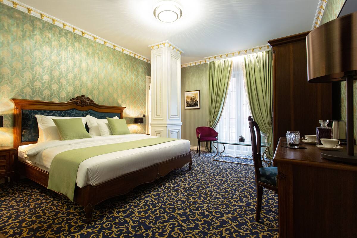 A hotel room with a large bed, a wooden nightstand, a desk with a chair, and a large window with curtains.