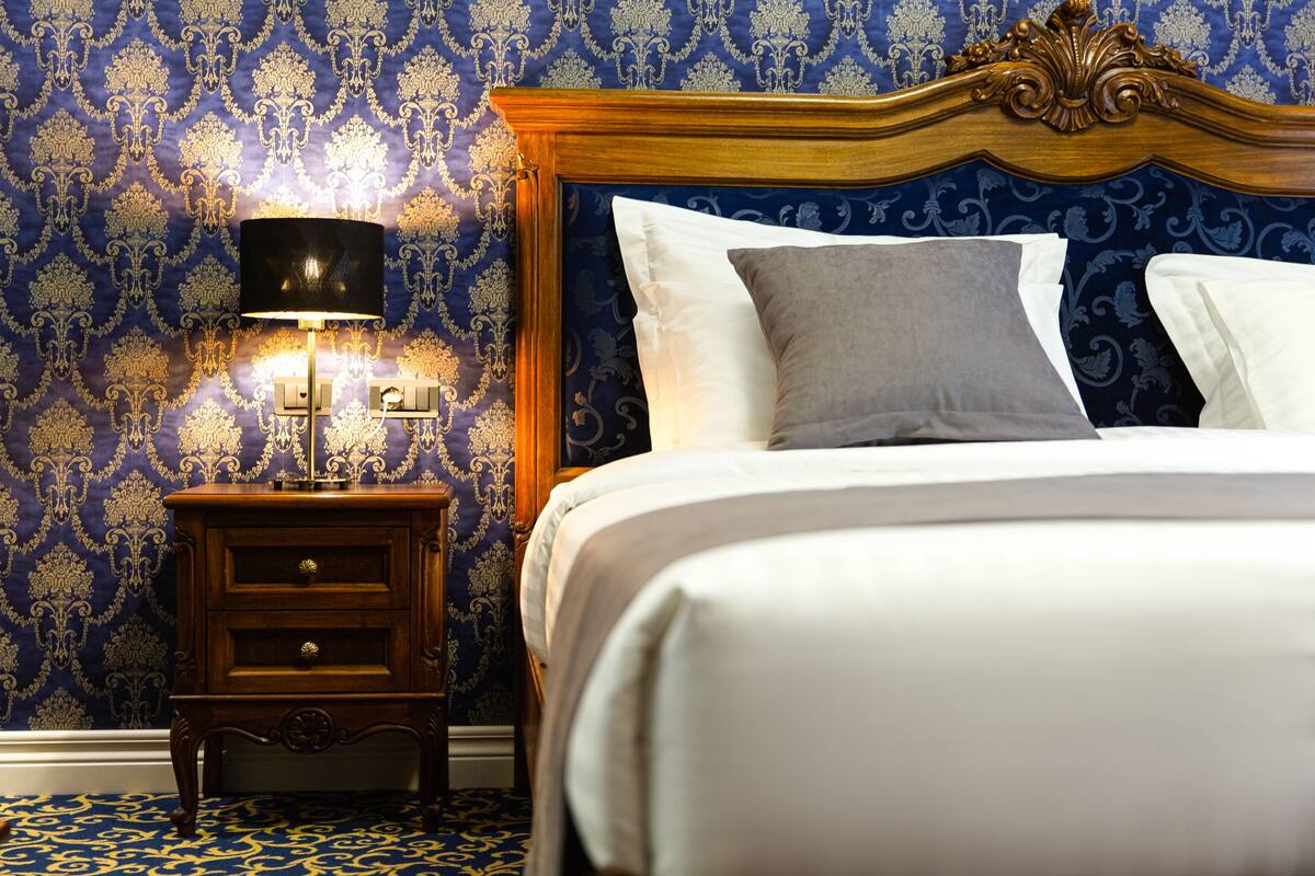A neatly made bed with white and gray linens, a wooden nightstand with a lamp, and a wall with a blue and gold damask pattern.