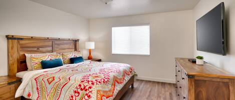 A bedroom with a wooden bed, a nightstand, a dresser, a TV, and a window with blinds.