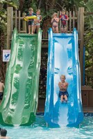 Un tobogán acuático con dos pistas separadas, una verde y otra azul, en un área de piscina rodeada de estructuras de madera y vegetación.
