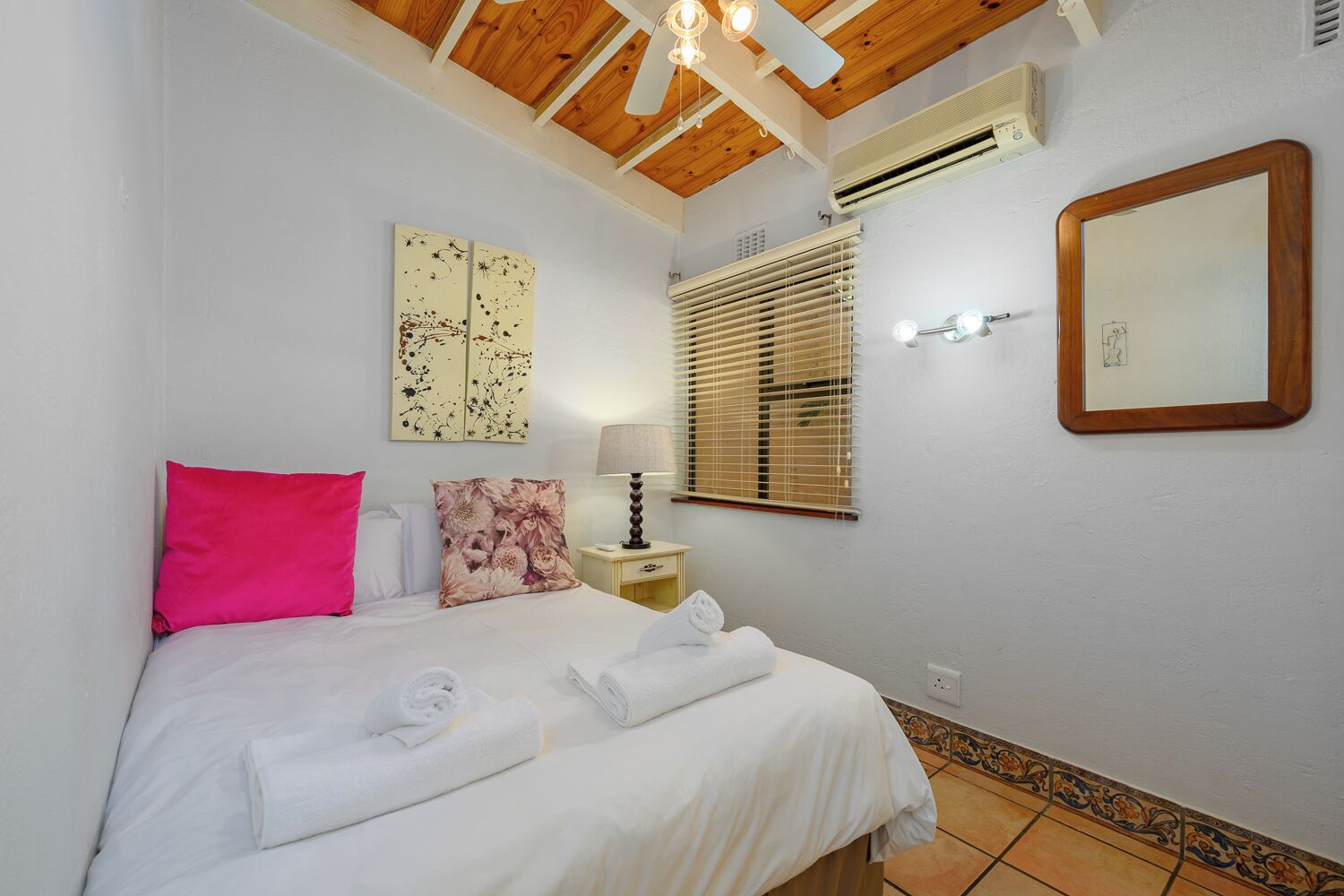 A bedroom with a bed, pillows, a nightstand, a lamp, a window with blinds, an air conditioning unit, a mirror, and artwork on the wall.