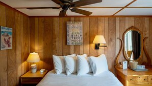 A hotel room with a wooden paneled wall, a bed with white bedding, two bedside lamps, a ceiling fan, a wooden dresser with a mirror, and a wall sign with various words. - Isleview Cottage 4 - 10 minutes to Acadia! (Trenton)