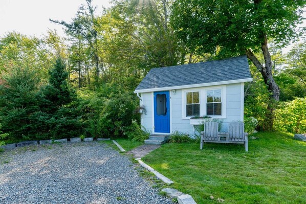 A small, single-story house with a blue door, surrounded by lush greenery and a gravel driveway. - Isleview Cottage 5 - 2 min to Mount Desert Isle! (Trenton)