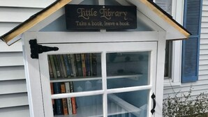Una piccola struttura bianca simile a una libreria con un cartello che dice "Little Library Take a book, leave a book". È posizionata su un pontile di legno con ringhiera, e sullo sfondo si vede una casa blu.