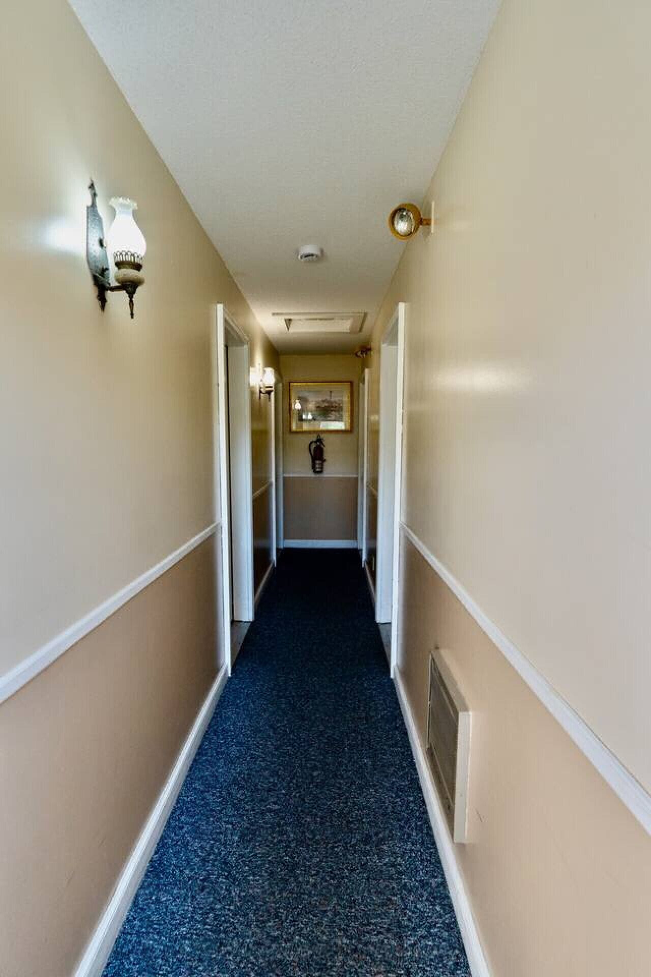 Un couloir étroit aux murs de couleur beige, un tapis bleu foncé et des moulures blanches. Deux appliques murales sont fixées de part et d’autre.