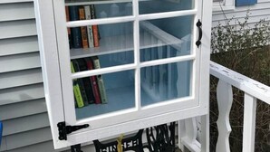 A small, white bookcase with a 'Take a book, leave a book' sign, placed on a wooden deck.